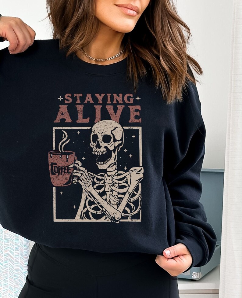 K&ouml;nnte beinhalten: Schwarzes Sweatshirt mit einer Grafik eines Skeletts, das eine Kaffeetasse h&auml;lt. Der Text "STAYING ALIVE" steht &uuml;ber dem Skelett. Auf der Kaffeetasse steht das Wort "COFFEE". Das Design befindet sich in einem rechteckigen Rahmen mit Sternen.