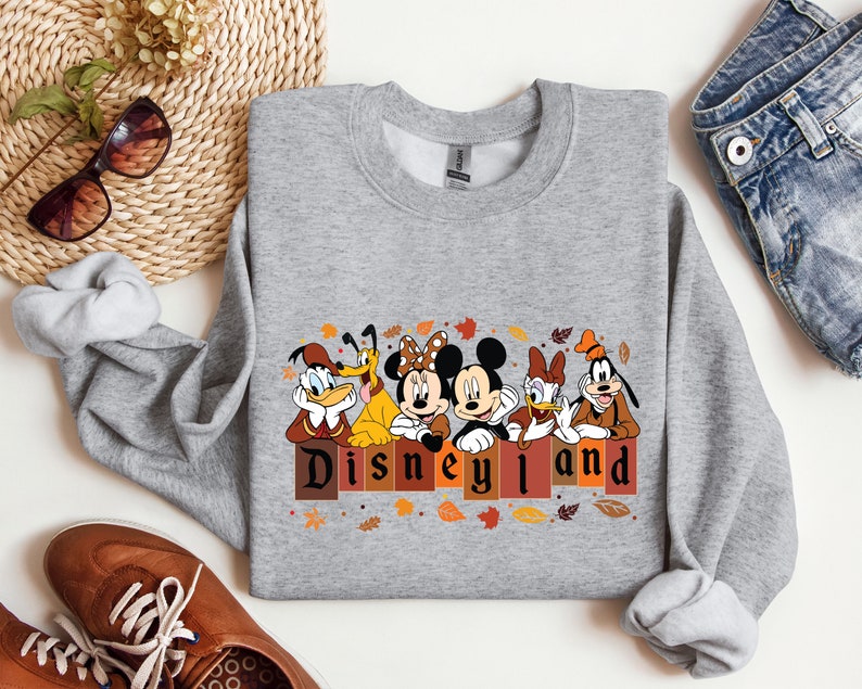 May include: Gray sweatshirt with a graphic of Mickey Mouse, Minnie Mouse, Donald Duck, Goofy, and Pluto in front of a brick wall with the text "Disneyland" in brown letters. The characters are wearing fall-themed outfits and there are fall leaves around them.