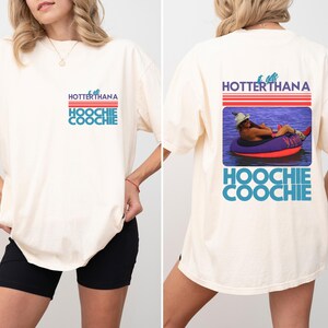May include: Cream-colored t-shirt with the text "HOTTERTHANA HOOCHIE COOCHIE" in red, blue, and white. The back of the shirt features a graphic of a person in a tube on the water. The shirt is a casual, oversized fit.