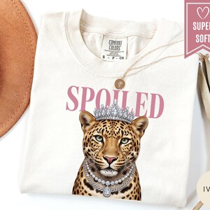 May include: Ivory-colored t-shirt with the word "SPOILED" in pink letters. The shirt features a leopard wearing a silver crown and necklace. The shirt has a round neckline and short sleeves. The words "SUPER SOFT" are in a pink banner.