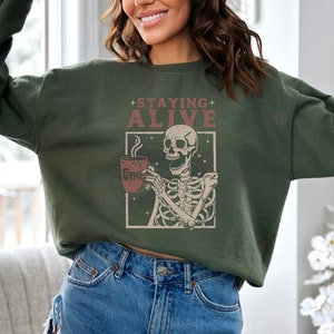K&ouml;nnte beinhalten: Dunkelgr&uuml;nes Sweatshirt mit dem Text "+ STAYING + ALIVE" &uuml;ber einer Skelett-Illustration, die eine Kaffeetasse h&auml;lt. Auf der Kaffeetasse steht das Wort "Coffee". Das Sweatshirt ist ein bequemer, l&auml;ssiger Stil.
