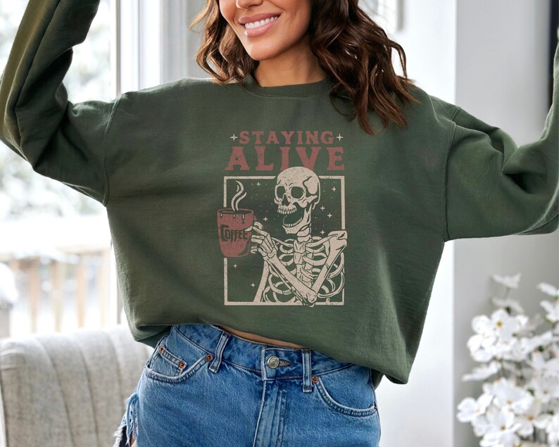 K&ouml;nnte beinhalten: Dunkelgr&uuml;nes Sweatshirt mit dem Text "+ STAYING + ALIVE" &uuml;ber einer Skelett-Illustration, die eine Kaffeetasse h&auml;lt. Auf der Kaffeetasse steht das Wort "Coffee". Das Sweatshirt ist ein bequemer, l&auml;ssiger Stil.