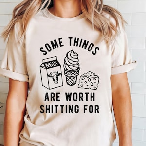 May include: A beige t-shirt with black text that reads "Some things are worth shitting for". The text is surrounded by a cartoon image of a carton of milk, an ice cream cone, and a slice of cheese.