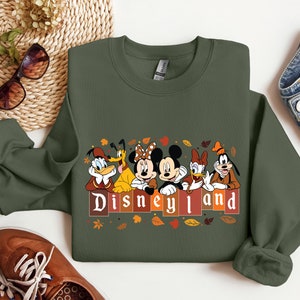 May include: A green sweatshirt with a print of Mickey Mouse, Donald Duck, Goofy, and Pluto in front of a sign that says "Disneyland". The characters are wearing fall-themed outfits and there are leaves scattered around them.