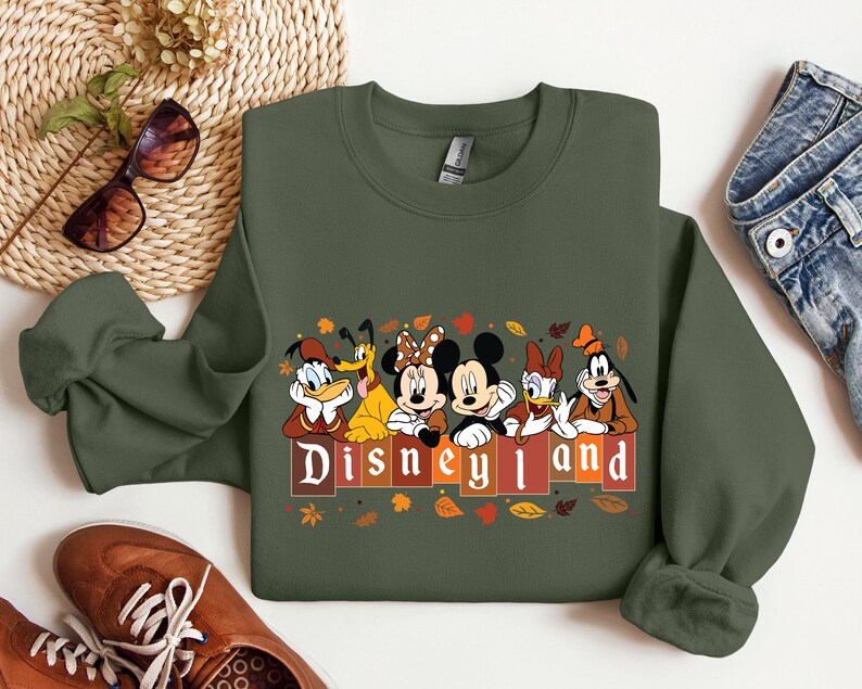May include: A green sweatshirt with a print of Mickey Mouse, Donald Duck, Goofy, and Pluto in front of a sign that says "Disneyland". The characters are wearing fall-themed outfits and there are leaves scattered around them.