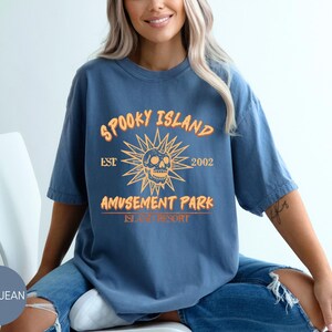 May include: Blue jean t-shirt with the text "Spooky Island Amusement Park" and a skull graphic. The design is in orange and yellow. The shirt is a short-sleeved, casual style.