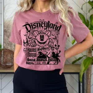 Puede incluir: Camiseta de manga corta color malva con un gr&aacute;fico negro. El gr&aacute;fico incluye la palabra "Disneyland" sobre un dise&ntilde;o de calabaza de Mickey Mouse. El gr&aacute;fico tambi&eacute;n incluye "Haunted Mansion" y "Oogie Boogie Bash".