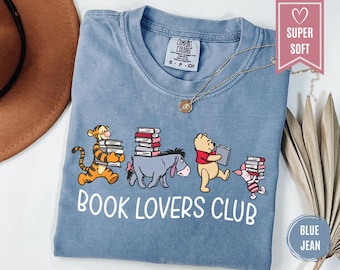 May include: A blue t-shirt featuring a graphic of Tigger, Eeyore, Winnie the Pooh, and Piglet carrying stacks of books. The text "BOOK LOVERS CLUB" is printed in white below the characters. The shirt has a crew neckline and a size small label.