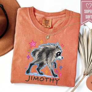 Puede incluir: Una camiseta de color terracota con una ilustraci&oacute;n gr&aacute;fica de un mapache gris caminando. El dise&ntilde;o incluye estrellas coloridas dibujadas a mano y el nombre JIMOTHY impreso en negro debajo del mapache. Etiqueta de la marca Comfort Colors.
