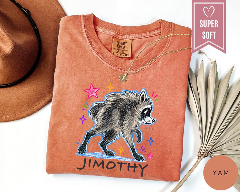 Puede incluir: Una camiseta de color terracota con una ilustraci&oacute;n gr&aacute;fica de un mapache gris caminando. El dise&ntilde;o incluye estrellas coloridas dibujadas a mano y el nombre JIMOTHY impreso en negro debajo del mapache. Etiqueta de la marca Comfort Colors.