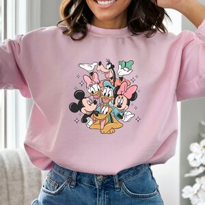 K&ouml;nnte beinhalten: Rosafarbener Pullover mit einem Aufdruck von Mickey Mouse, Minnie Mouse, Donald Duck, Goofy und Pluto. Die Figuren sind in einem verspielten Stapel angeordnet, mit Mickey Mouse unten und Goofy oben.