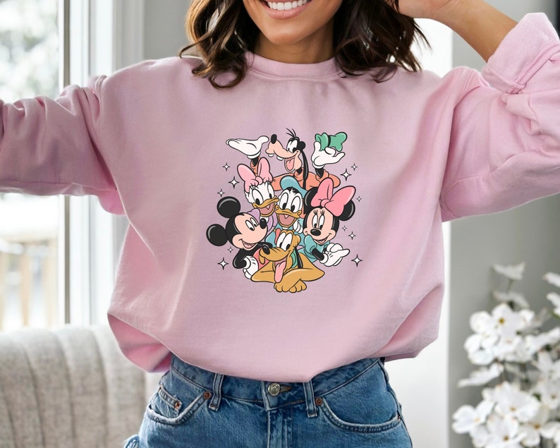 K&ouml;nnte beinhalten: Rosafarbener Pullover mit einem Aufdruck von Mickey Mouse, Minnie Mouse, Donald Duck, Goofy und Pluto. Die Figuren sind in einem verspielten Stapel angeordnet, mit Mickey Mouse unten und Goofy oben.