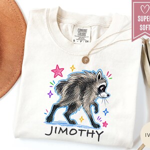 Puede incluir: Una camiseta de color marfil con una ilustraci&oacute;n gr&aacute;fica de un mapache de pelaje oscuro y cara blanca. El mapache est&aacute; rodeado de estrellas y destellos coloridos dibujados a mano. Debajo del mapache, el nombre JIMOTHY est&aacute; impreso en negro.