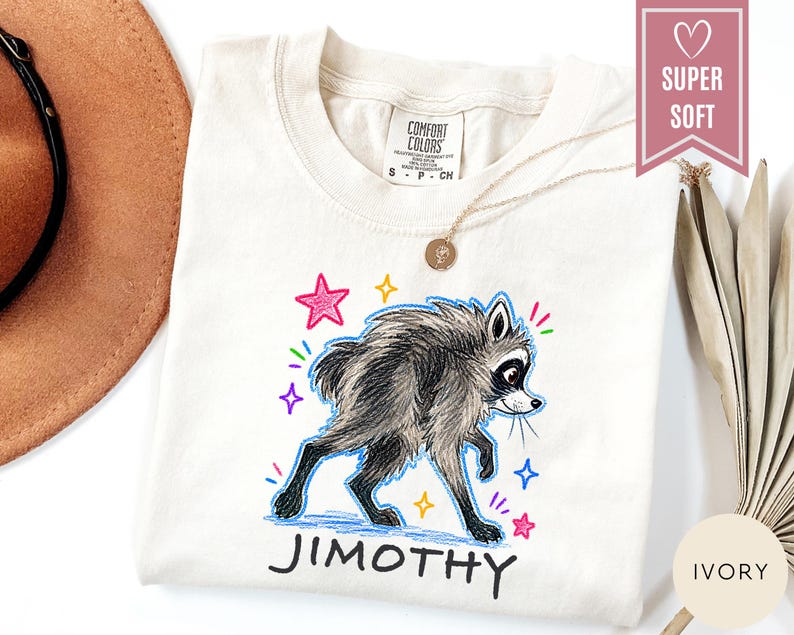 Puede incluir: Una camiseta de color marfil con una ilustraci&oacute;n gr&aacute;fica de un mapache de pelaje oscuro y cara blanca. El mapache est&aacute; rodeado de estrellas y destellos coloridos dibujados a mano. Debajo del mapache, el nombre JIMOTHY est&aacute; impreso en negro.