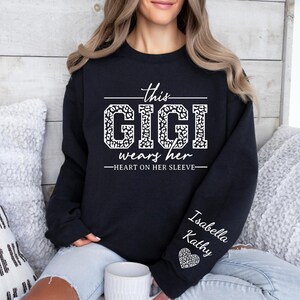 May include: Black sweatshirt with white text that reads "This Gigi wears her heart on her sleeve". The text is surrounded by a leopard print design. The sleeve has the names "Isabella" and "Kathy" written in white text.