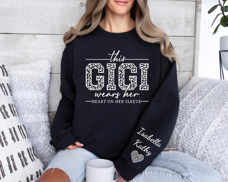 May include: Black sweatshirt with white text that reads "This Gigi wears her heart on her sleeve". The text is surrounded by a leopard print design. The sleeve has the names "Isabella" and "Kathy" written in white text.