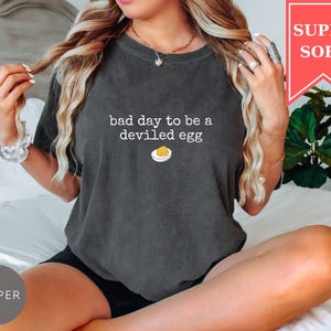 May include: Dark grey t-shirt with the text "bad day to be a deviled egg" and a graphic of a deviled egg. The shirt has a relaxed fit and is labeled "SUPER SOFT" in a red banner.