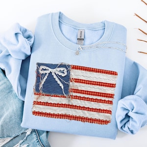 May include: A light blue sweatshirt with a decorative American flag design. The flag is made of fabric with red and white stripes and a blue field with a white bow. A silver necklace with a pendant is also visible.