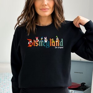 Puede incluir: Sudadera negra con un logotipo de Disneyland colorido que presenta a Mickey Mouse, Minnie Mouse, Donald Duck, Goofy y Pluto. El texto "The Original" est&aacute; debajo del logotipo.