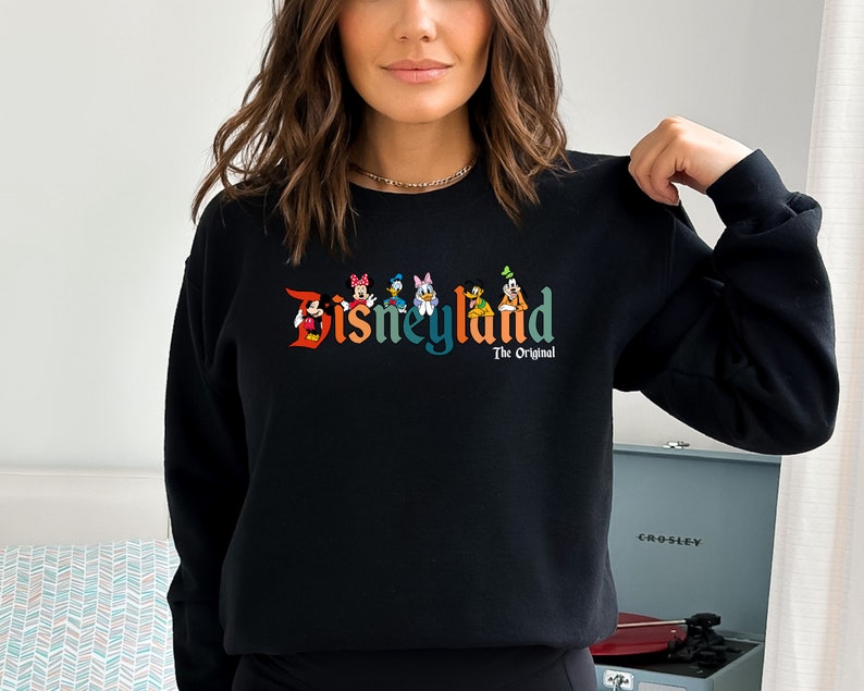 Puede incluir: Sudadera negra con un logotipo de Disneyland colorido que presenta a Mickey Mouse, Minnie Mouse, Donald Duck, Goofy y Pluto. El texto "The Original" est&aacute; debajo del logotipo.