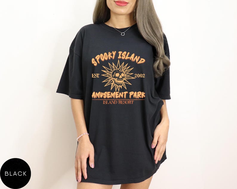 May include: Black t-shirt with the text "SPOOKY ISLAND AMUSEMENT PARK ISLAND RESORT" in orange. The design includes a skull, sunburst, and the year "2002". The shirt is a casual, oversized fit.