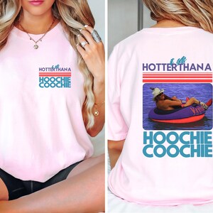May include: Light pink t-shirt with the text "Hotter Than a Hoochie Coochie" in red, white, and blue. The back of the shirt features a graphic of a person in a tube on the water with the same text.