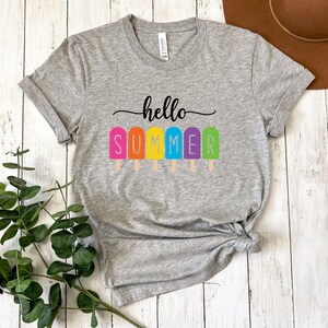 May include: A heather gray t-shirt with the words "hello SUMMER" printed in black script above colorful popsicle graphics. The popsicles are pink, orange, yellow, blue, and green. The shirt is knotted at the waist.