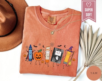 Può includere: Una t-shirt color ruggine con una stampa grafica di cinque articoli di cancelleria animati vestiti per Halloween: un pastello vampiro, una zucca, una colla stick mummia, un quaderno con un fantasma e una matita strega con scopa.