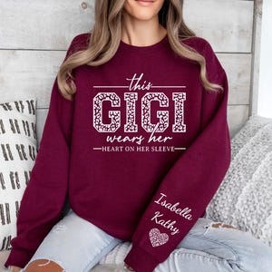 May include: Burgundy sweatshirt with white text that reads "This Gigi wears her heart on her sleeve". The text is in a leopard print font. The sweatshirt has a white heart with the names "Isabella Kathy" on the sleeve.