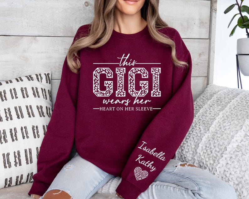 May include: Burgundy sweatshirt with white text that reads "This Gigi wears her heart on her sleeve". The text is in a leopard print font. The sweatshirt has a white heart with the names "Isabella Kathy" on the sleeve.