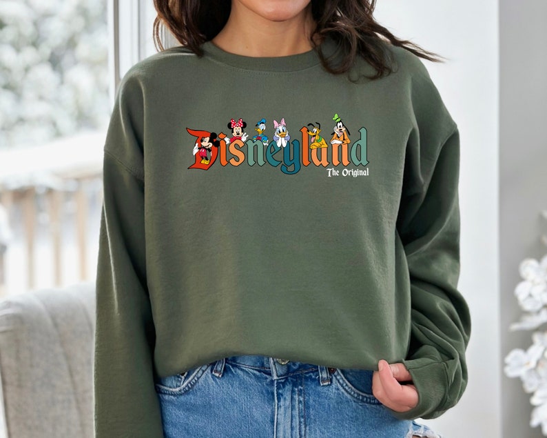 Puede incluir: Una sudadera verde con un logotipo de Disneyland de estilo vintage que presenta a Mickey Mouse, Minnie Mouse, Donald Duck, Goofy y Pluto. El texto "The Original" est&aacute; impreso debajo del logotipo.