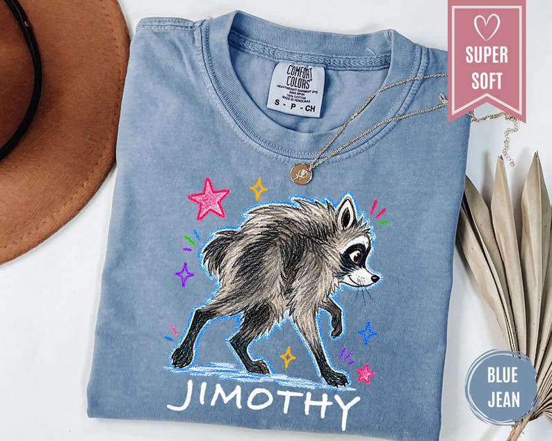 Puede incluir: Una camiseta de manga corta color azul gris&aacute;ceo con una ilustraci&oacute;n gr&aacute;fica de un mapache de pelaje esponjoso. El dise&ntilde;o incluye estrellas coloridas dibujadas a mano y el nombre JIMOTHY impreso en letras may&uacute;sculas blancas bajo el gr&aacute;fico.
