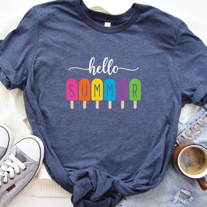 May include: A navy blue t-shirt with the words "hello SUMMER" in white script and colorful popsicle graphics. The shirt is paired with gray sneakers, denim shorts, and a cup of coffee.