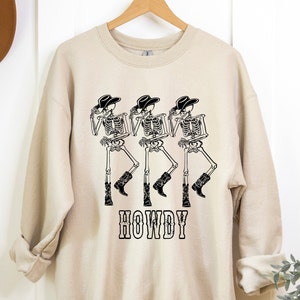 May include: A beige sweatshirt with a graphic of three skeletons wearing cowboy hats and boots. The skeletons are dancing and the text "HOWDY" is printed below them.