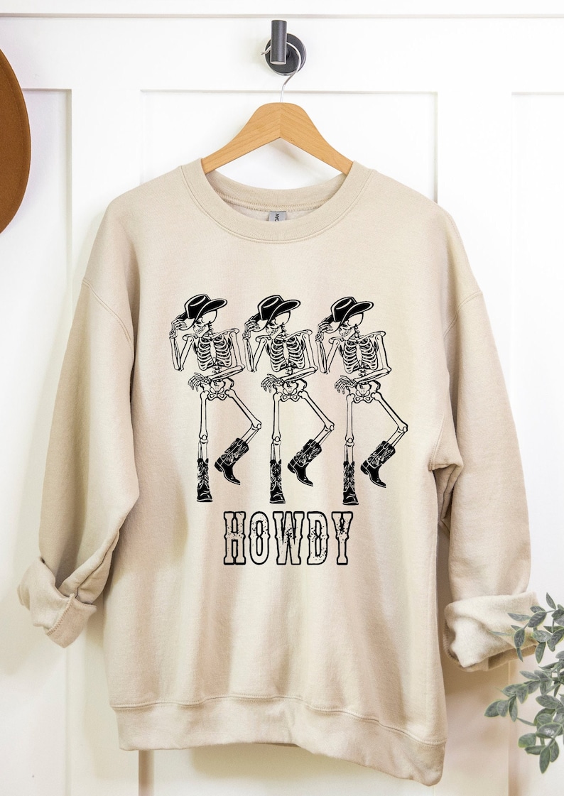 May include: A beige sweatshirt with a graphic of three skeletons wearing cowboy hats and boots. The skeletons are dancing and the text "HOWDY" is printed below them.