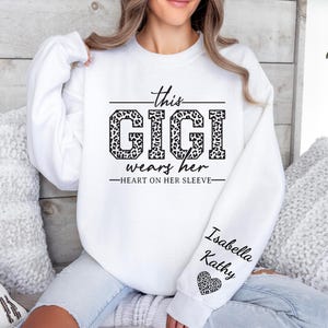 May include: White sweatshirt with the text "This Gigi wears her heart on her sleeve" and a leopard print design. The sleeve has the names "Isabella" and "Kathy" written on it with a leopard print heart.