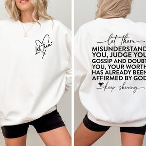 May include: White sweatshirt with a black heart and the text "Let Them" on the front and the text "Let them MISUNDERSTAND YOU, JUDGE YOU, GOSSIP AND DOUBT YOU, YOUR WORTH HAS ALREADY BEEN AFFIRMED BY GOD. keep shining" on the back.