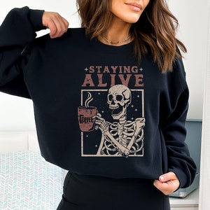 K&ouml;nnte beinhalten: Schwarzes Sweatshirt mit einer Grafik eines Skeletts, das eine Kaffeetasse h&auml;lt. Der Text "STAYING ALIVE" steht &uuml;ber dem Skelett. Auf der Kaffeetasse steht das Wort "Coffee". Das Design ist im Vintage-Stil.