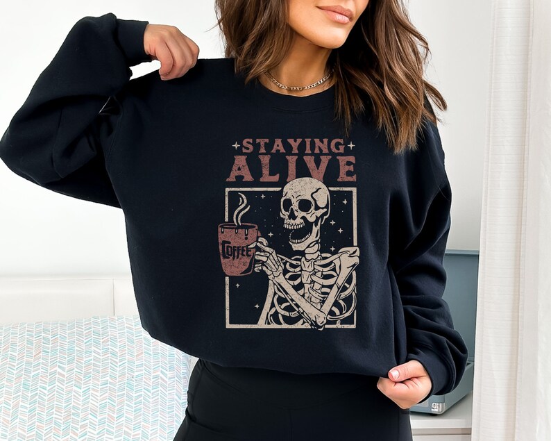 K&ouml;nnte beinhalten: Schwarzes Sweatshirt mit einer Grafik eines Skeletts, das eine Kaffeetasse h&auml;lt. Der Text "STAYING ALIVE" steht &uuml;ber dem Skelett. Auf der Kaffeetasse steht das Wort "Coffee". Das Design ist im Vintage-Stil.