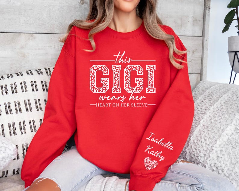 May include: Red sweatshirt with white text that reads "This Gigi wears her heart on her sleeve" with a leopard print design. The sleeve has the names "Isabella Kathy" and a white heart.