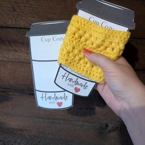 Pode incluir: Uma capa de crochê para copo em amarelo brilhante com um padrão de pontos texturizados. A capa é exibida num cartão de papel branco em forma de copo de café, com o texto impresso Cup Cozy e Handmade com um pequeno ícone de coração vermelho.
