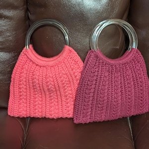 May include: Two handmade, knitted handbags. One is a vibrant coral color, the other is a deep magenta. Both have clear, circular handles. The bags are made with a textured stitch pattern, giving them a unique, handcrafted look.