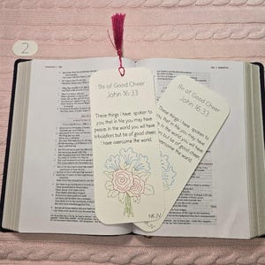 May include: Open Bible with two decorative bookmarks. The bookmarks are white with a floral design and the text "Be of Good Cheer John 16:33". A pink tassel is attached to the top of one bookmark.