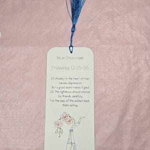 May include: A white bookmark with a blue tassel, featuring the text "Be an Encourager" and a Bible verse from Proverbs 12:25-26. The bookmark includes a small illustration of flowers in a vase. The letters "NKJV" are at the bottom.