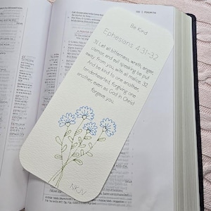 May include: A white bookmark with a floral design and text from Ephesians 4:31-32. The bookmark features a bouquet of blue embroidered flowers and the words "Be Kind" at the top. The bookmark is placed in an open book.