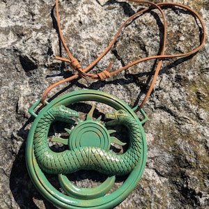 Peut inclure: Pendentif vert orné de deux serpents se faisant face, encerclant un motif solaire. Il est suspendu à un cordon en cuir marron. Le pendentif repose sur une surface en pierre texturée.