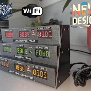 Può includere: Un orologio digitale in stile retrò con tre pannelli che mostrano l'ora e la data. L'orologio ha una finitura metallica grigia con display a LED rossi, verdi e gialli. Il testo "NEW DESIGN" è visibile. Ha anche un simbolo Wi-Fi.