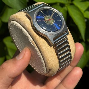 May include: A vintage wristwatch with a blue face and a sunburst design in gold and red. The watch has a silver-tone metal band and case. The watch is displayed on a beige watch pillow.