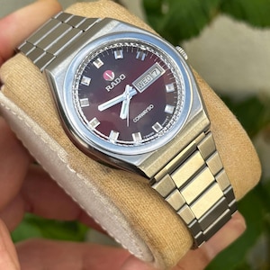 May include: A silver Rado Conway 50 wristwatch with a burgundy dial. The watch has a day-date display and a silver link bracelet. The watch is displayed on a tan surface.