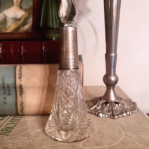 May include: A clear glass perfume bottle with a faceted crystal stopper and a silver collar. The glass body features a diamond-cut pattern, while the silver neck displays small engraved hallmarks. The bottle has a wide, tapered base.